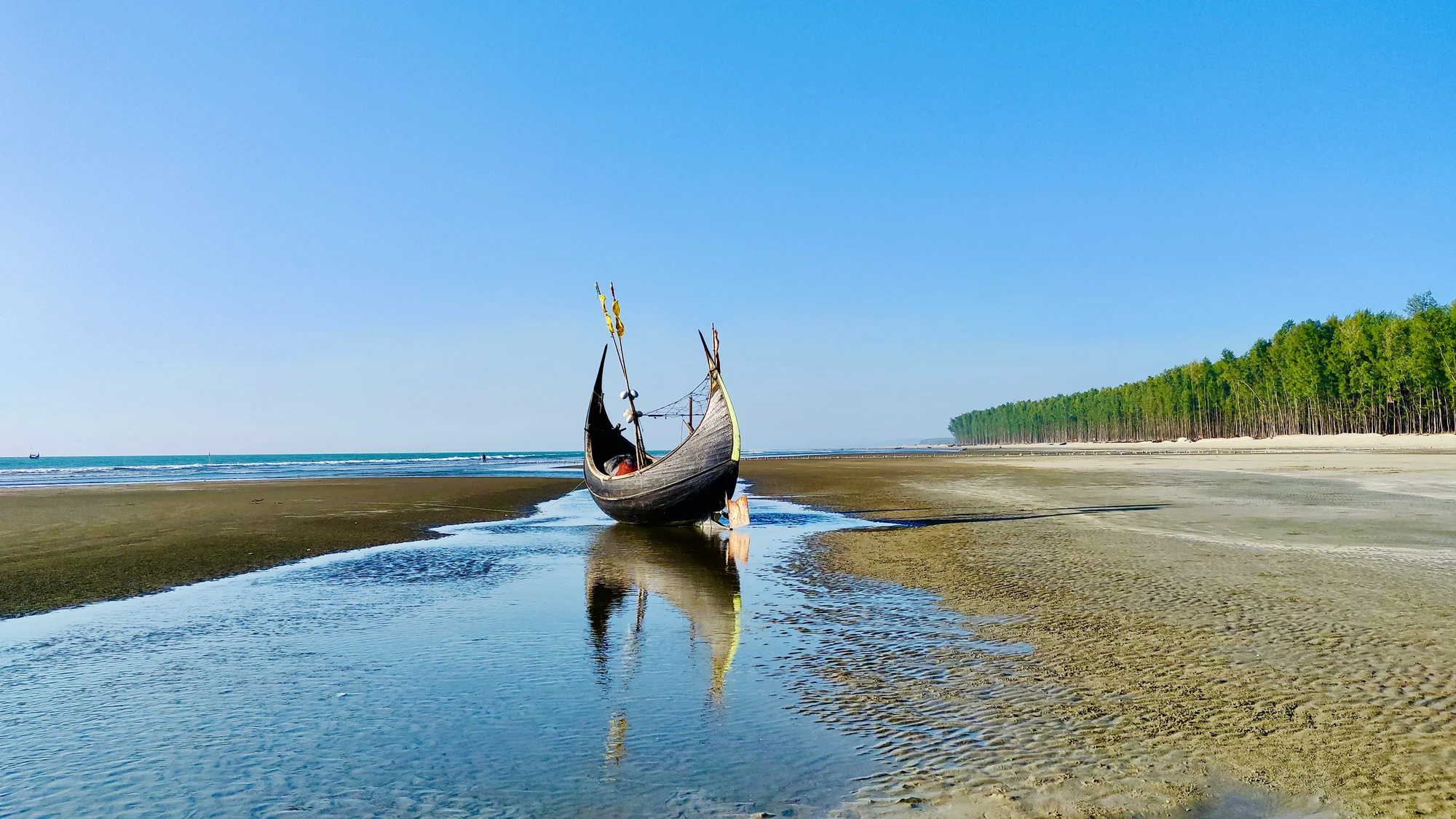 Traditional boat on Cox's Bazar beach with clear sky and tree line.