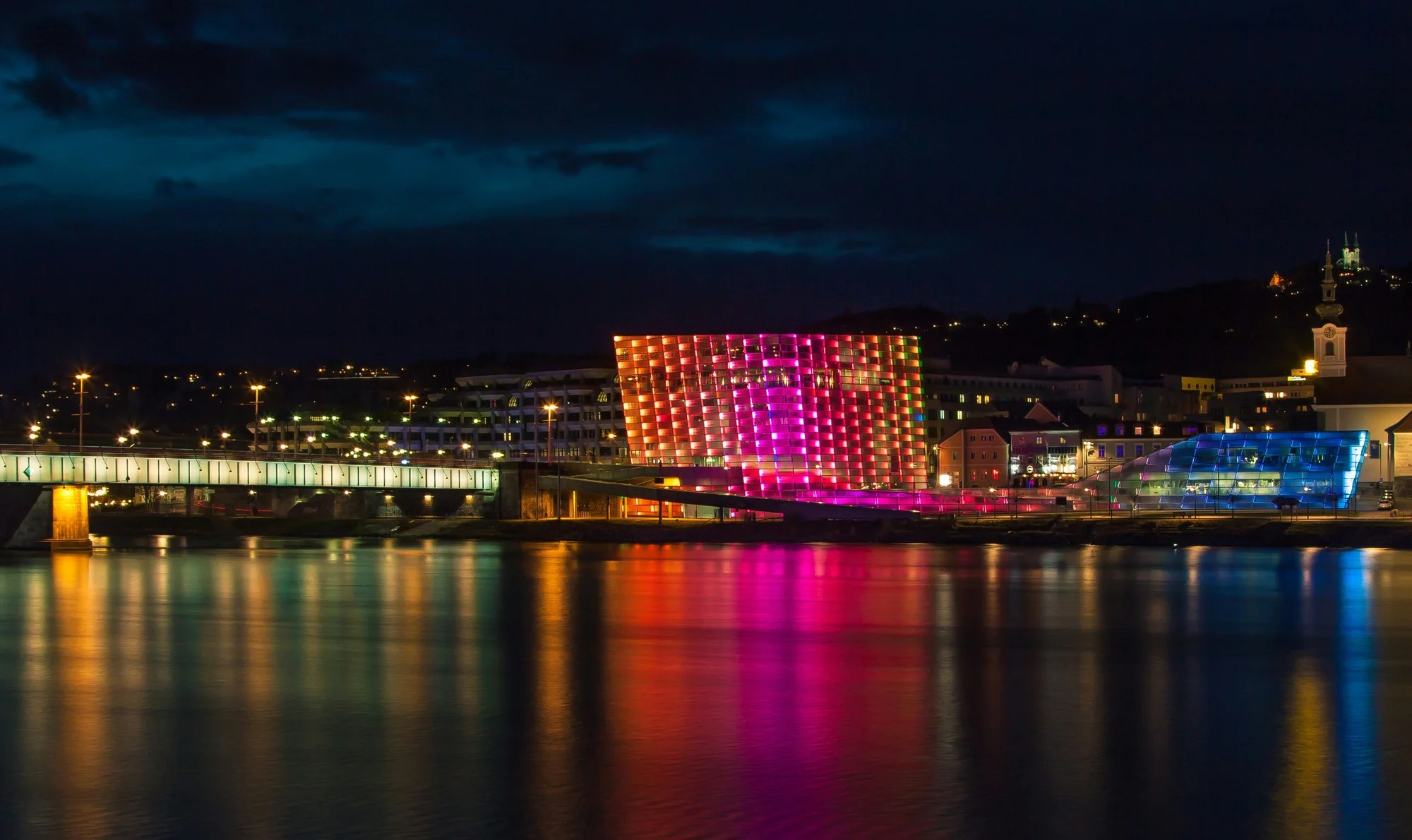 Ars Electronica Center and Nibelungen Bridge at night in Linz, with Danube reflecting colorful lights.