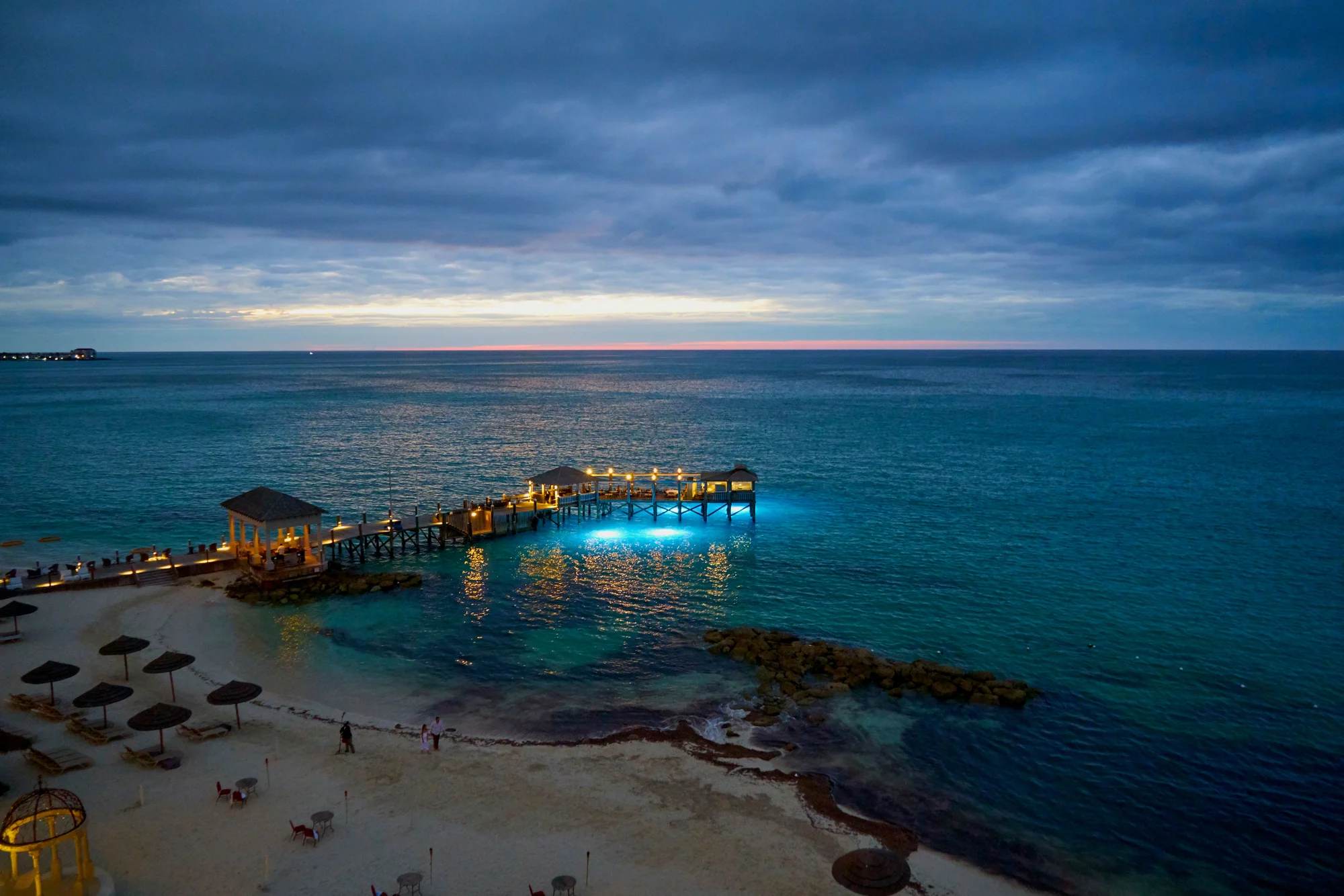 Beach sunset in Nassau, Bahamas, with a lit pier and turquoise ocean.