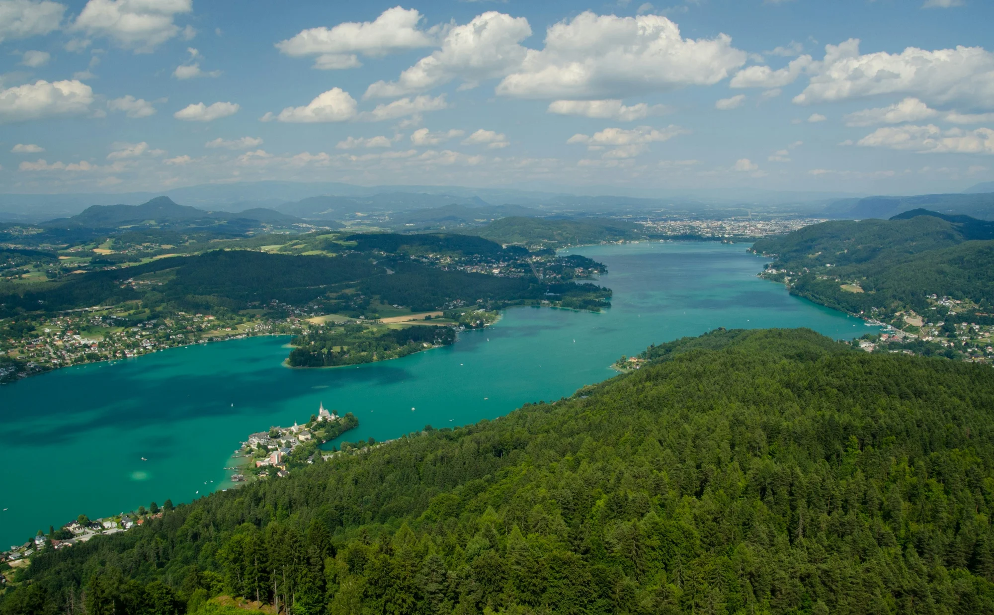 Aerial view of a lake in Klagenfurt, Austria, surrounded by green hills and dense forests.