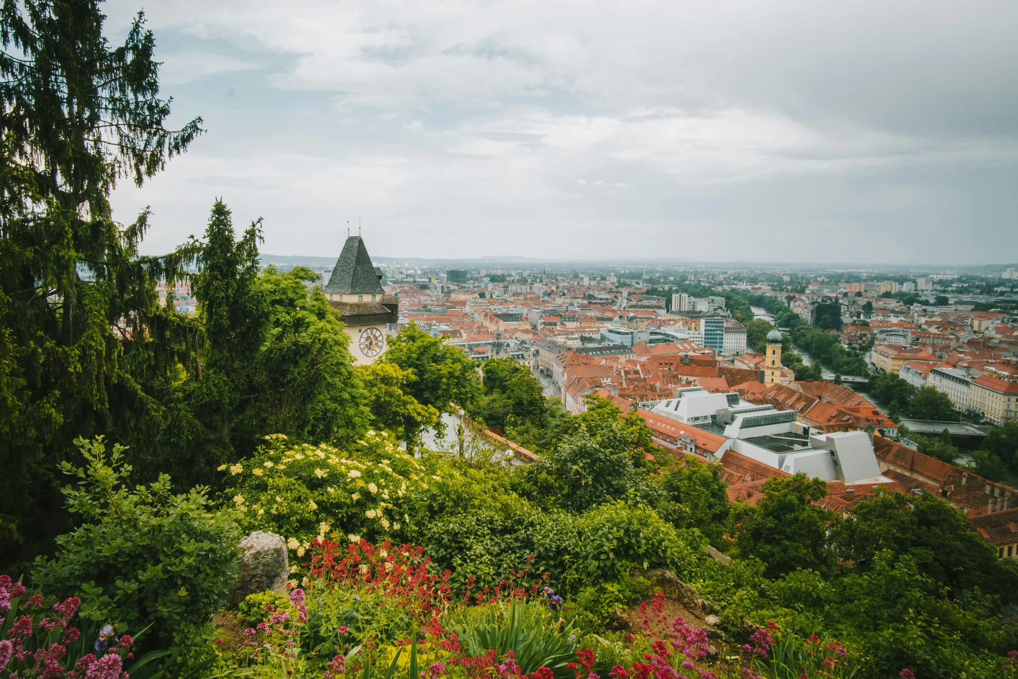 Clock Tower of Graz, Austria, surrounded by trees and flowers with the cityscape in the background.