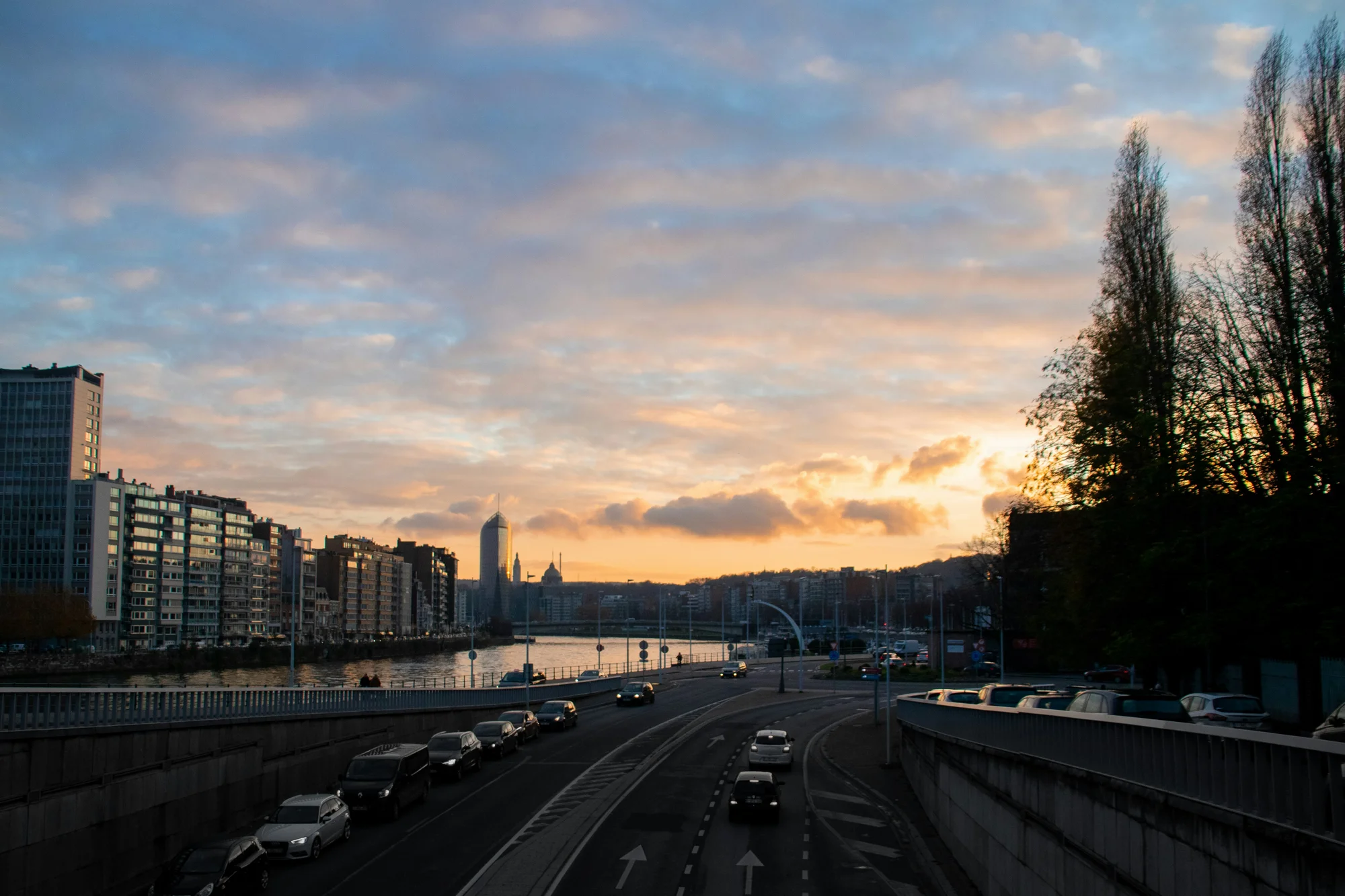 Sunset over the Meuse River in Liège, Belgium, with modern buildings and cars on the road.